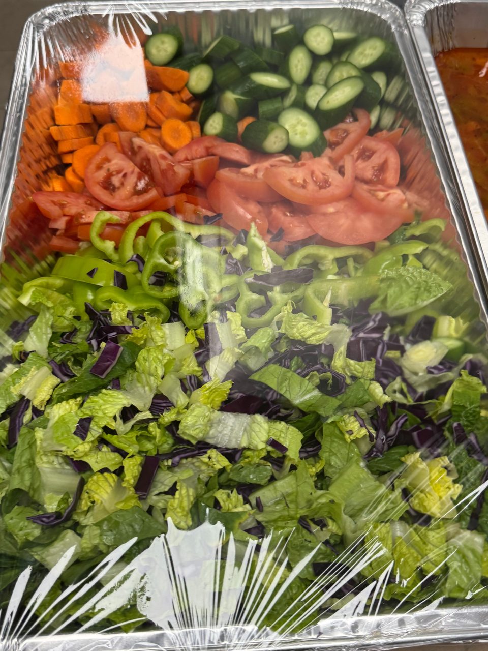 A foil tray filled with fresh salad ingredients, including lettuce, bell peppers, tomatoes, carrots, and cucumbers.