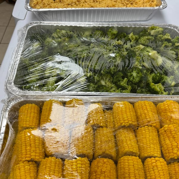 Three foil trays of food, including rice, broccoli, and corn on the cob, covered with plastic wrap.