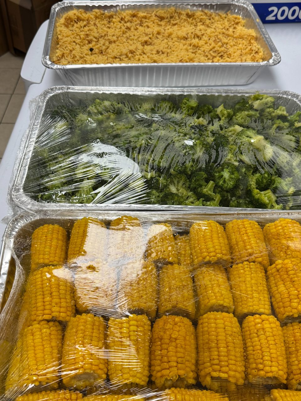 Three foil trays of food, including rice, broccoli, and corn on the cob, covered with plastic wrap.