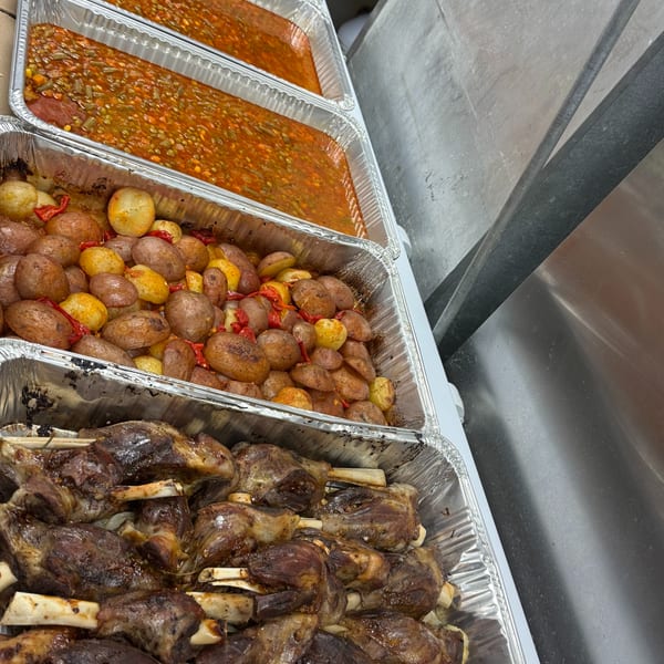 Four aluminum trays filled with cooked lamb shanks, roasted potatoes, vegetable stew, and fresh salad.
