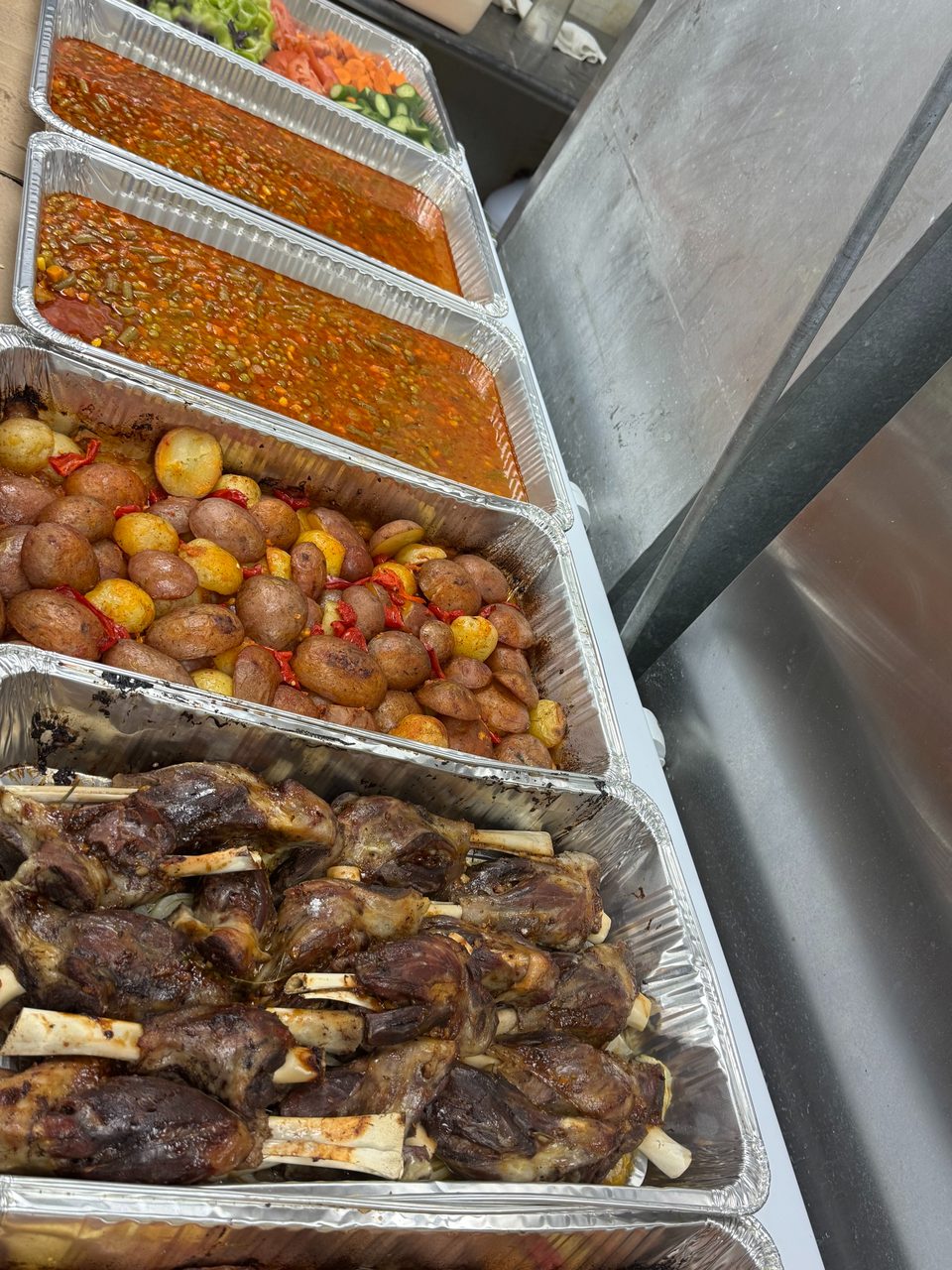 Four aluminum trays filled with cooked lamb shanks, roasted potatoes, vegetable stew, and fresh salad.