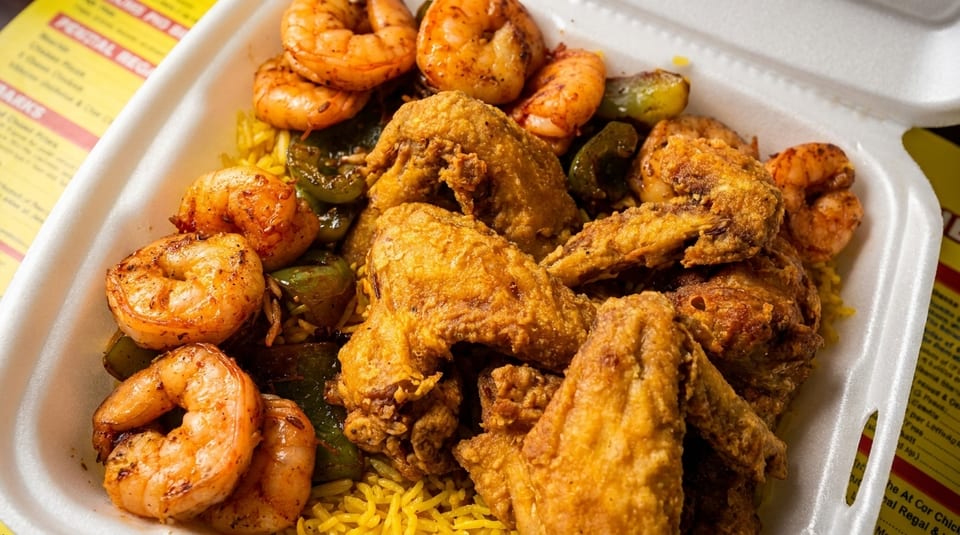 Fried chicken wings and grilled shrimp with green peppers over yellow rice in a white takeout container.