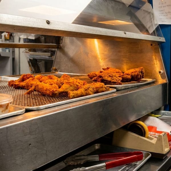 Crispy fried chicken pieces on metal trays under a warming lamp in a restaurant kitchen.